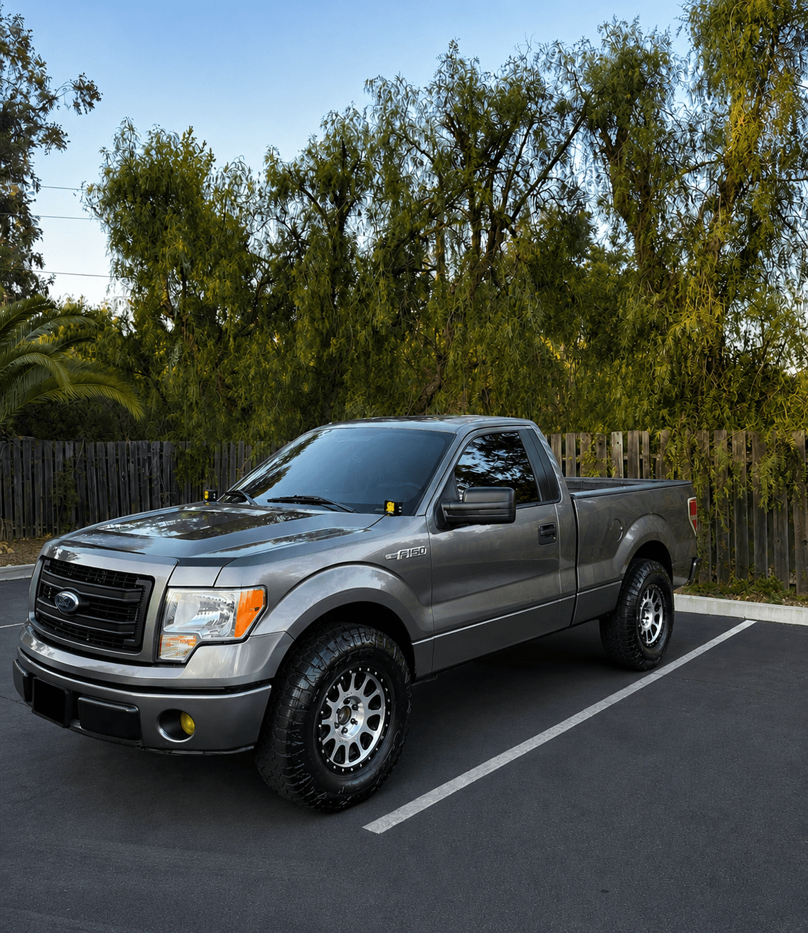 Charcoal metallic Ford F-150 regular cab pickup with aftermarket wheels and all-terrain tires, parked in a lot at golden hour with warm light on the glossy exterior