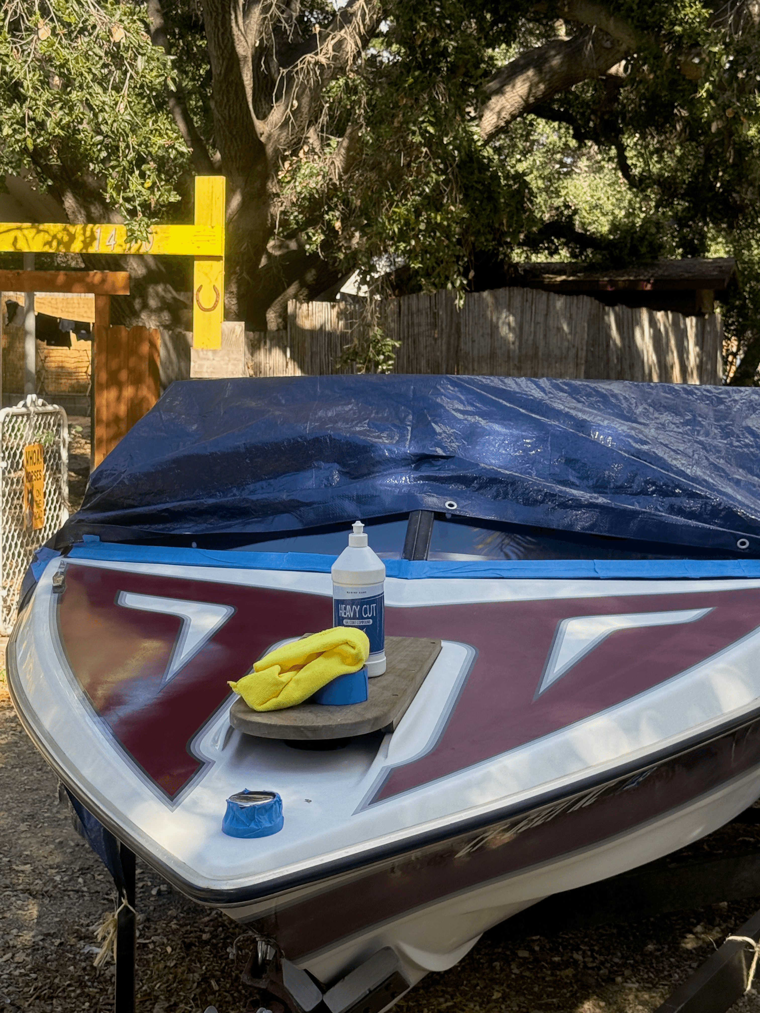 White and maroon motorboat bow on a trailer with heavy-cut compound and microfiber towels during gelcoat polishing